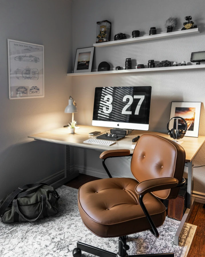 A designer's home studio with a flip-clock iMac, leather chair, and shelves of cameras